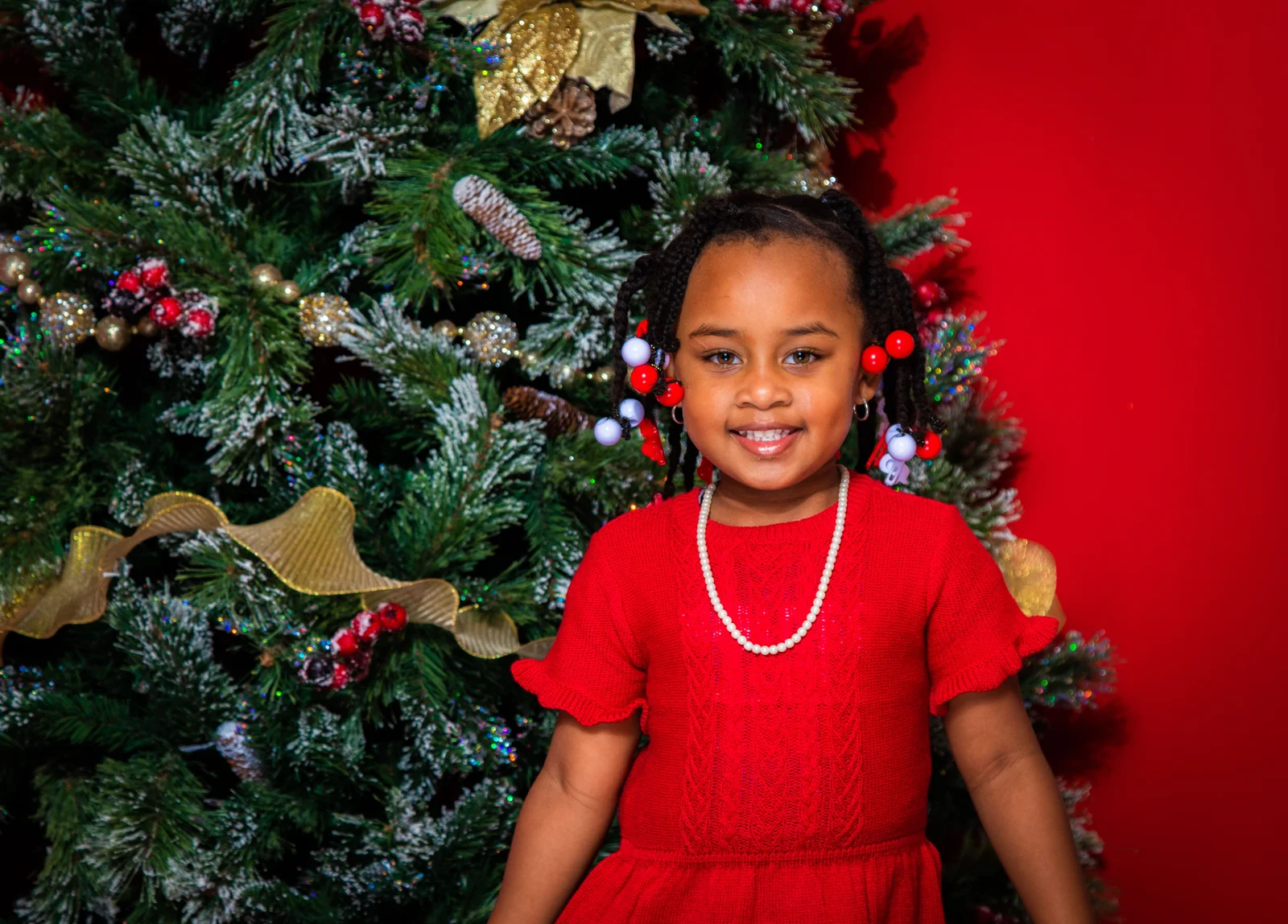 Petite fille en robe de fête rouge et collier de perles devant le sapin de Noël