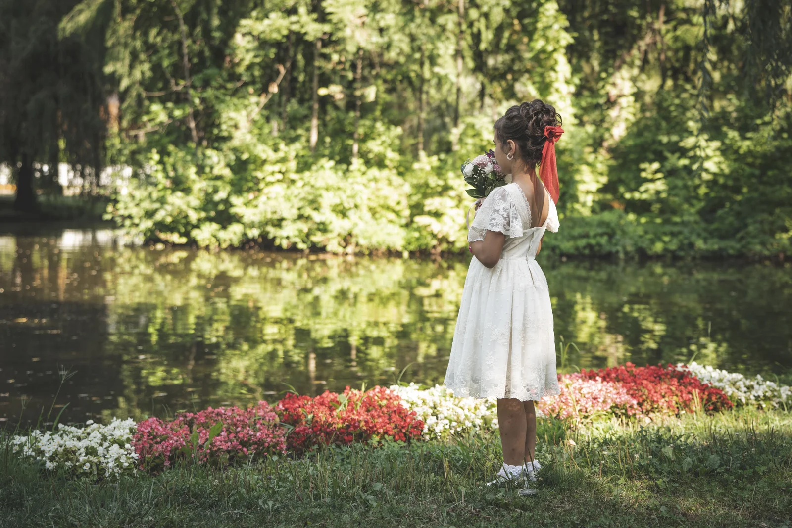 Fille en robe de cérémonie blanche vue de dos au bord d'un bassin fleuri